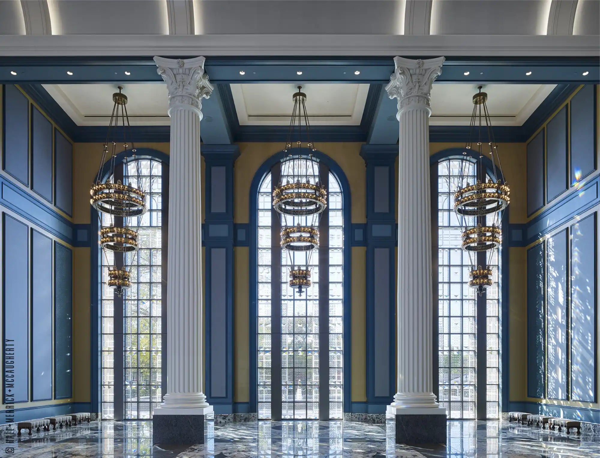 A grand hall with tall arched windows, large chandeliers, and decorative columns.