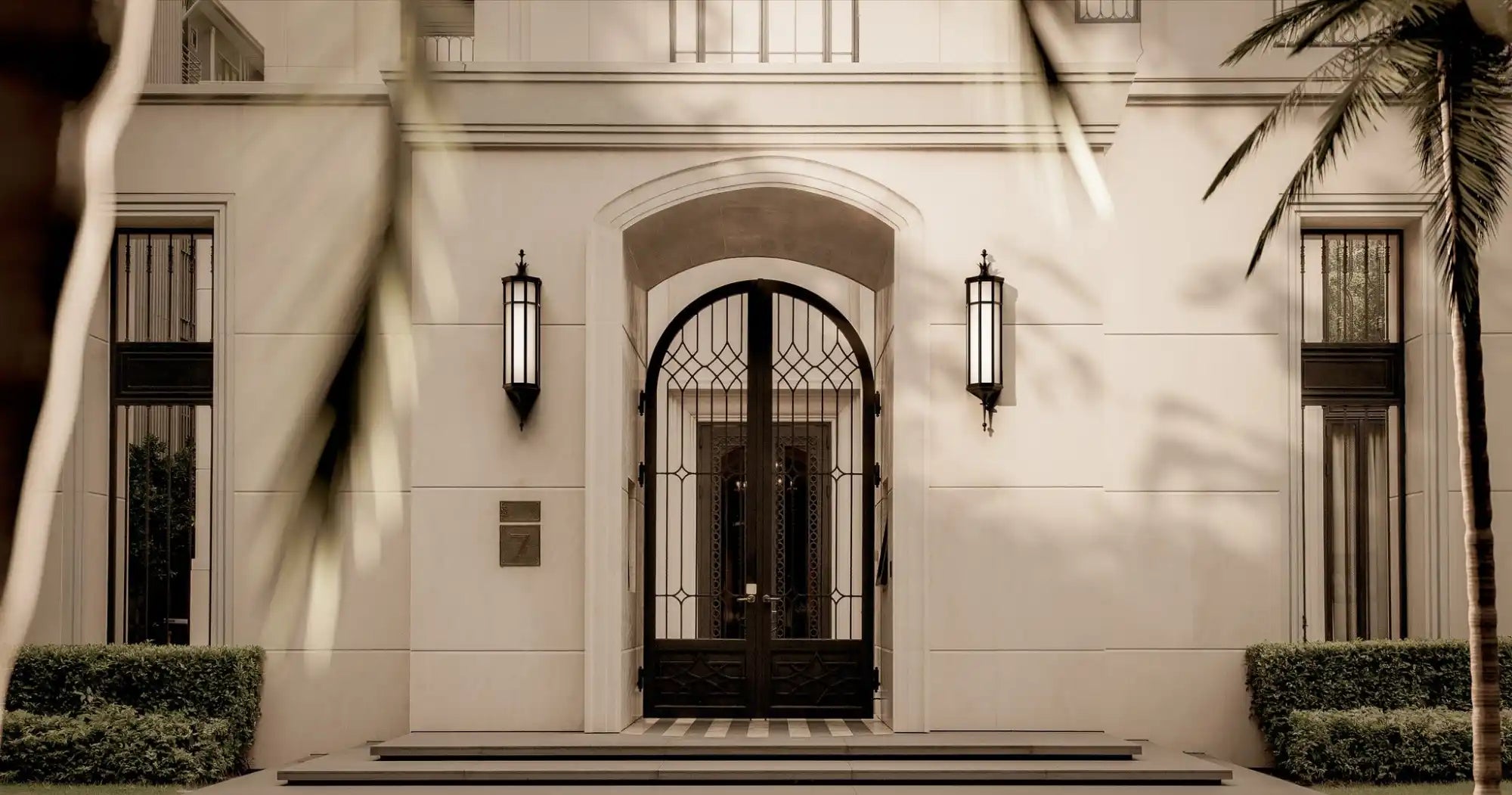 A grand, light-colored building facade with a prominent arched entryway featuring an ornate black iron gate, flanked by tall windows and elegant wall sconces.