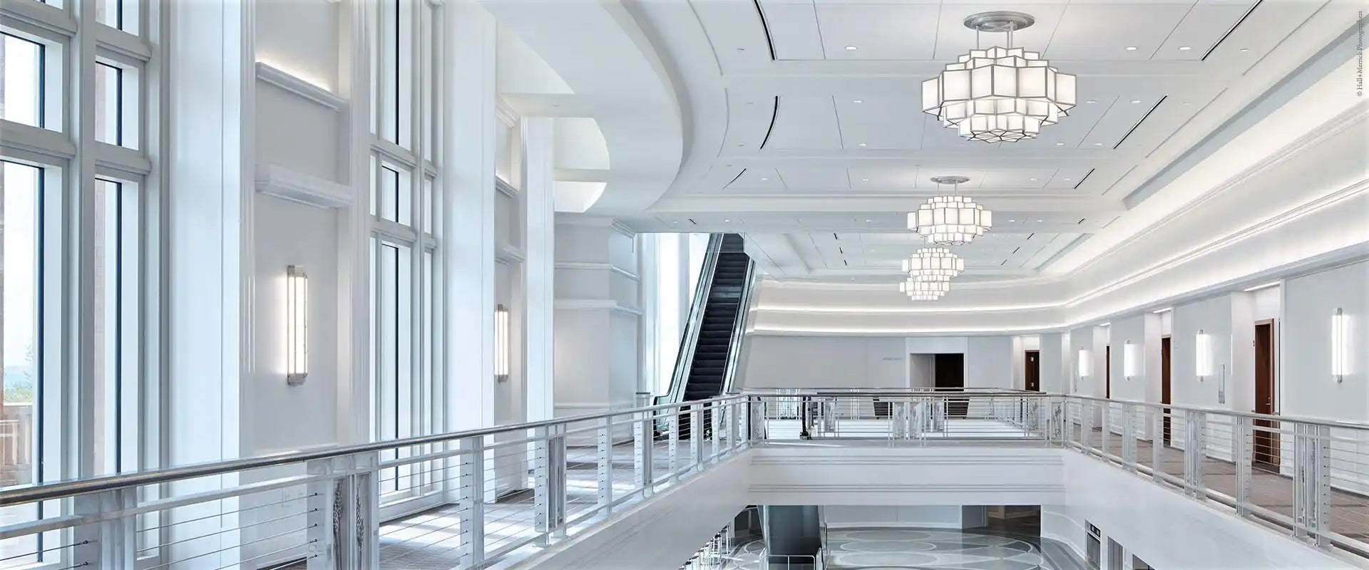 A modern, multi-level interior with white walls, large windows, an escalator, and decorative chandeliers.