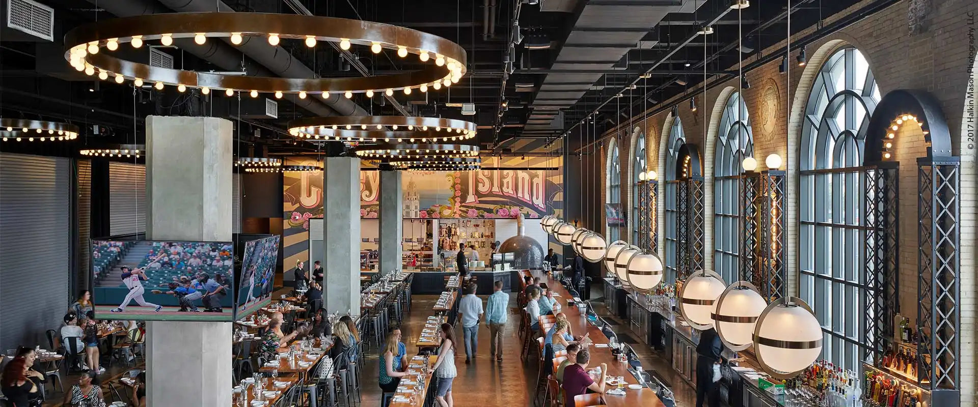 A spacious, modern restaurant or bar with high ceilings, long communal tables, arched windows, and decorative lighting.