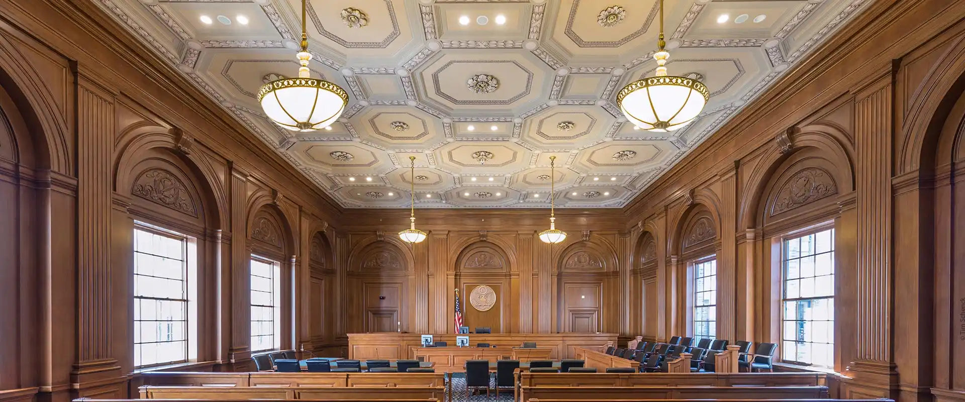 An ornate, wood-paneled courtroom with a coffered ceiling and large windows.